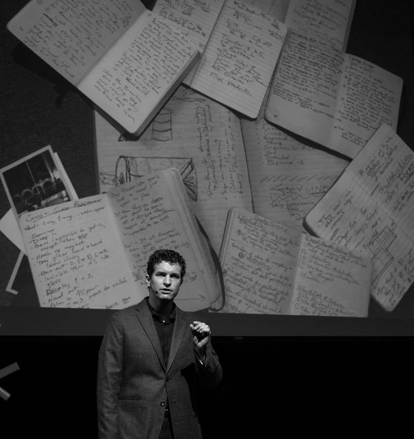 Evan Northrup on stage with handwritten notebooks projected behind him, black and white